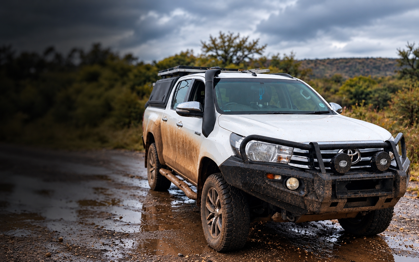 Muddy white Toyota Hilux with bullbar, snorkel and canopy on a wet gravel road under overcast skies